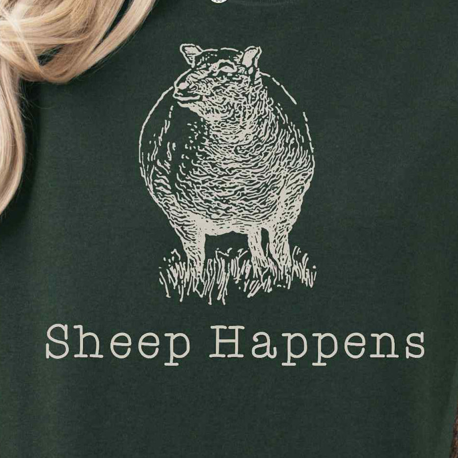 Person wearing a dark green t-shirt with 'Sheep Happens' text and graphic, sitting in a cozy indoor setting.